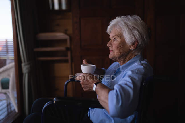 Vista lateral de una mujer mayor activa discapacitada reflexiva sosteniendo una taza de café y sentada en una silla de ruedas en el dormitorio en casa - foto de stock
