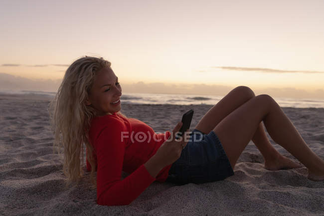 Вид сбоку на блондинку, которая пользуется мобильным телефоном, лежа на пляже. Она лежит на песке. — стоковое фото