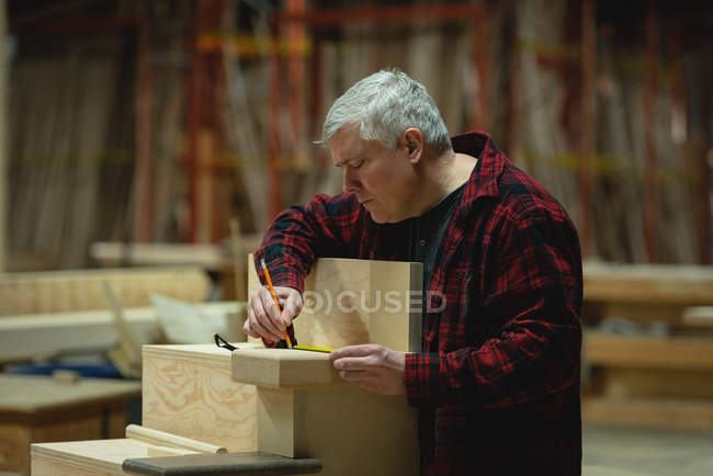 Vue de face du menuisier mesurant la planche de bois avec ruban à mesurer à l'atelier — Photo de stock