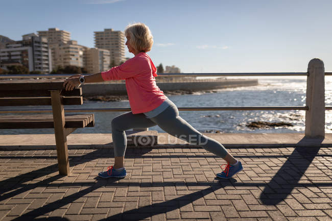 Seitenansicht einer aktiven Seniorin, die sich auf einer Strandpromenade in der Sonne gegen eine Bank streckt — Stockfoto