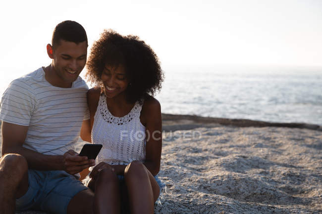 Vue de face du couple afro-américain utilisant un téléphone portable à la plage — Photo de stock