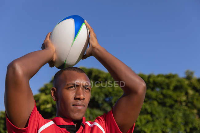 Vista frontal de un jugador de rugby afroamericano sosteniendo la pelota sobre su cabeza para ponerse en contacto en el estadio en un día soleado - foto de stock