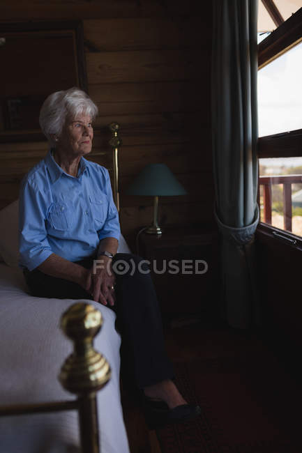Вид збоку на вдумливу активну старшу жінку, яка дивиться через вікно, сидячи на ліжку вдома — стокове фото