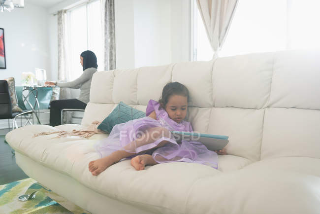 Вид спереди дочери смешанной расы, использующей цифровой планшет, в то время как мать в хиджабе работает на ноутбуке на заднем плане в гостиной дома — стоковое фото