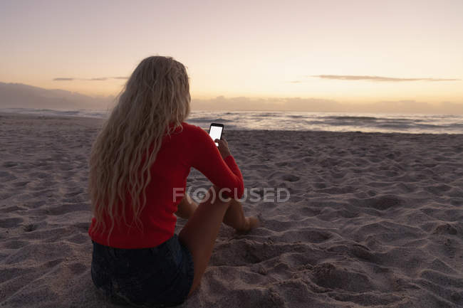 Vue arrière de la femme caucasienne blonde utilisant un téléphone mobile à la plage. Elle est assise sur le sable — Photo de stock
