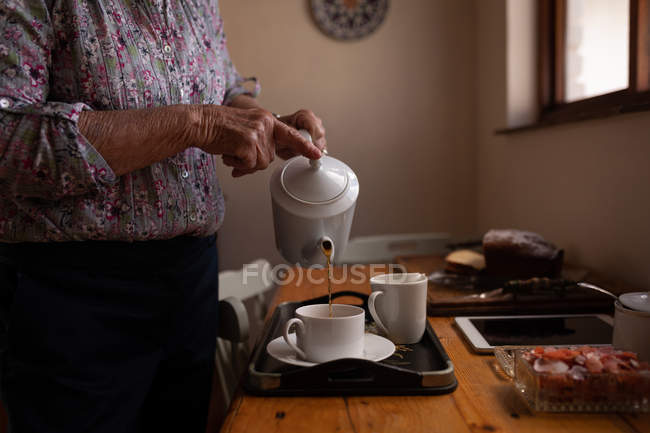 Partie médiane d'une femme âgée active versant du café dans une tasse à table dans la cuisine à la maison — Photo de stock