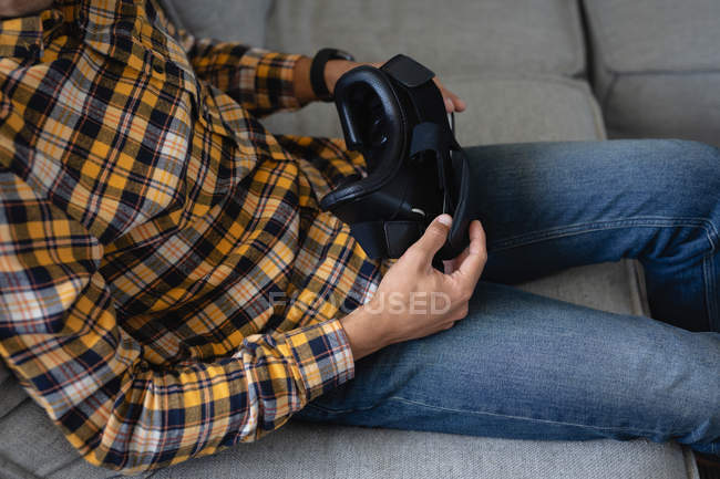 Середня частина людини, що тримає гарнітуру віртуальної реальності, сидить на дивані вдома — стокове фото