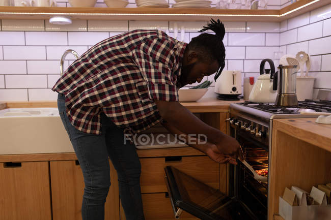 Vista lateral del hombre afroamericano insertando pizza en el horno en la cocina casera - foto de stock