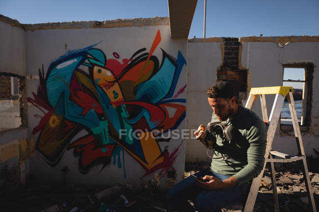 Front view of young Caucasian graffiti artist having food while using mobile phone — Stock Photo