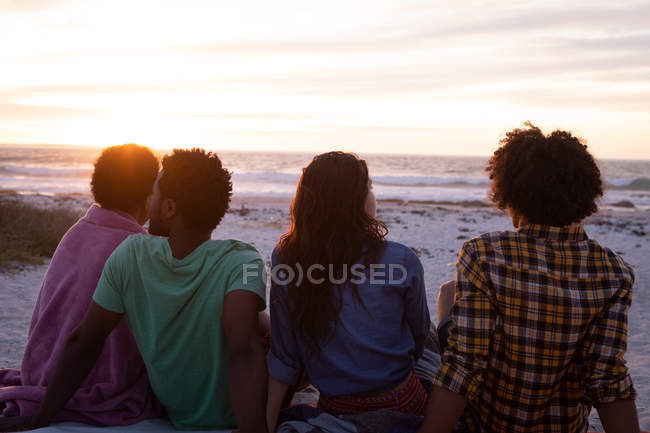 Вид спереду багатоетнічної групи друзів, що сидить на пляжі і дивиться далеко від океану під час заходу сонця — стокове фото