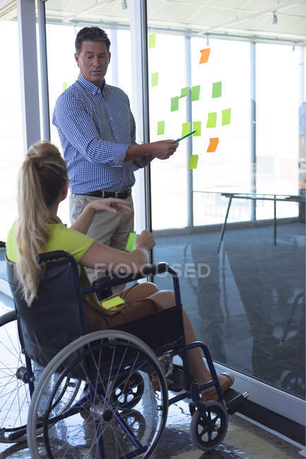 Vue arrière d'une femme blonde caucasienne handicapée et d'un dirigeant masculin caucasien discutant de notes collantes au bureau — Photo de stock