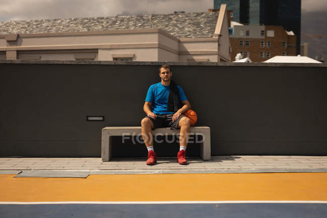 Retrato do jovem jogador caucasiano relaxando na quadra de basquete no banco contra a cidade em segundo plano. Ele está olhando para a câmera — Fotografia de Stock