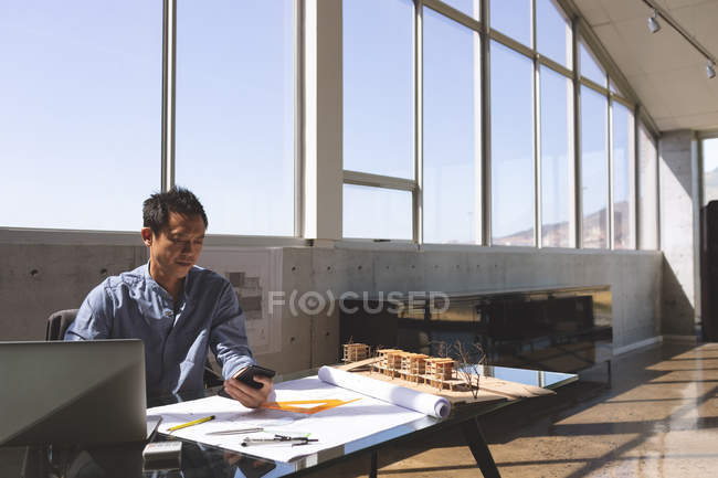 Vista frontal do arquiteto masculino asiático sentado na mesa enquanto usa telefone celular e modelo arquitetônico, régua triangular, lápis e planta na mesa no escritório moderno — Fotografia de Stock