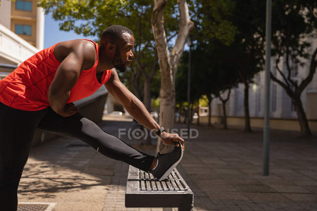Seitenansicht eines jungen afrikanisch-amerikanischen fitten Mannes, der an einem sonnigen Tag Dehnübungen auf einer Bank macht — Stockfoto