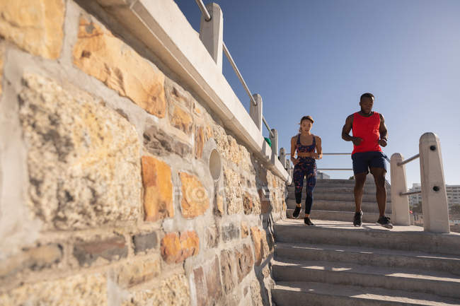 Вид спереду молода багатоетнічна пара біжить зі сходів біля пляжу в сонячний день — стокове фото