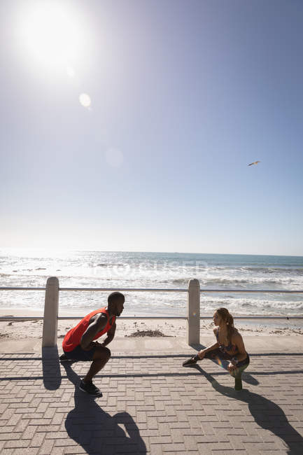 Front view of young multi-ethnic couple exercising on pavement near beach on a sunny day — Stock Photo