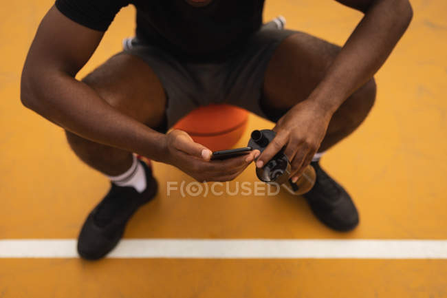 Niedriger Abschnitt des Spielers entspannt sich auf dem Basketballplatz, während er Mobiltelefon benutzt — Stockfoto