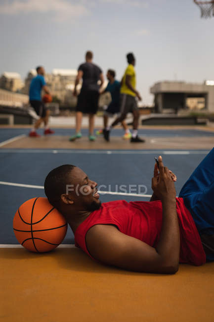 Vista lateral do feliz jogador de basquete afro-americano deitado no chão do parque infantil com a cabeça apoiada no basquete enquanto ele está usando o telefone celular no campo de basquete contra os jogadores que jogam no fundo — Fotografia de Stock