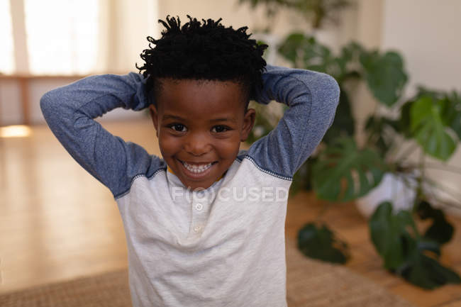 Retrato de niño afroamericano lindo feliz de pie con las manos detrás de la cabeza en casa. Él sonríe y mira a la cámara - foto de stock