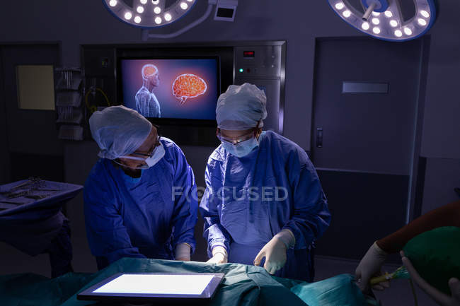 Front view of surgeons concentrated performing operation in operating room at hospital against spots and digital screen in background — Stock Photo