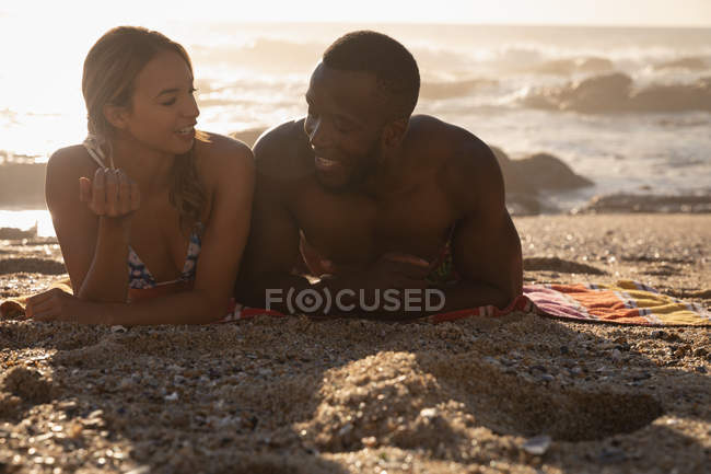 Vista frontal do jovem casal multi-étnico interagindo uns com os outros na praia em um dia ensolarado — Fotografia de Stock