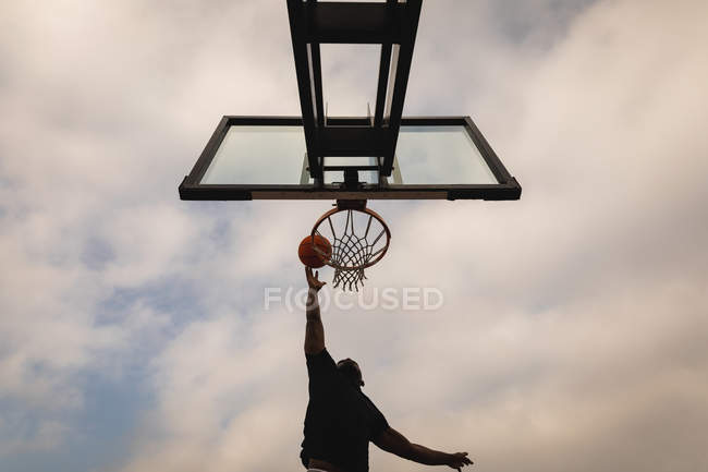 Vista de ángulo bajo del joven jugando baloncesto mientras se pone la pelota en el aro de baloncesto - foto de stock