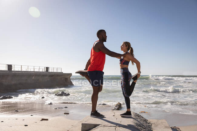 Vista lateral do jovem casal multi-étnico fazendo exercício de alongamento perto da praia em um dia ensolarado — Fotografia de Stock