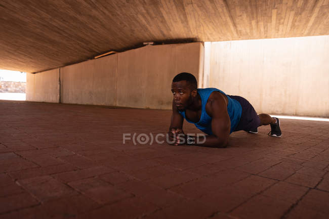 Vista lateral de un joven afroamericano en forma haciendo ejercicio de tablón bajo el puente en un día soleado - foto de stock