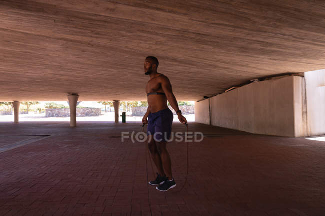 Side view of young African-American fit man doing skipping exercise with rope under the bridge — Stock Photo