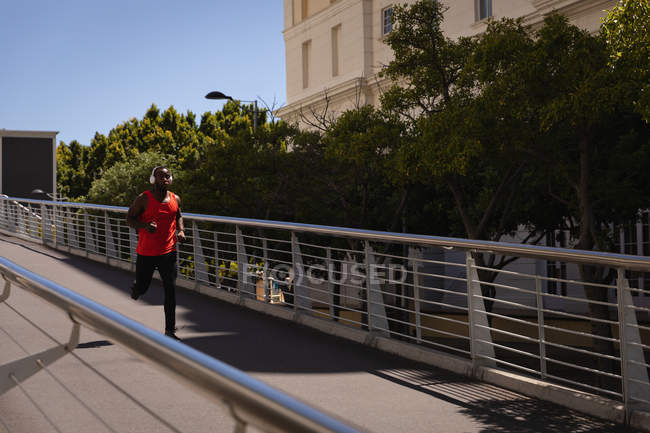 Frontansicht eines jungen afrikanisch-amerikanischen fitten Mannes mit Headset, der an einem sonnigen Tag gegen das Geländer läuft — Stockfoto