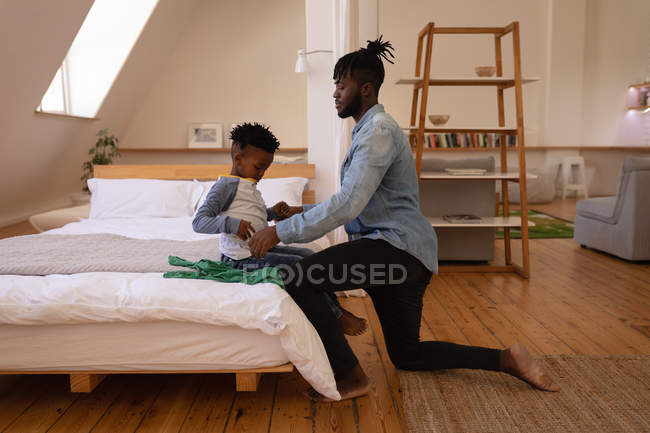 Vista lateral del padre afroamericano vistiendo hijo travieso en casa - foto de stock