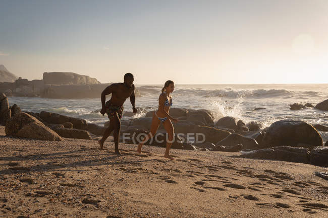 Vue latérale d'un jeune couple multi-ethnique courant du sable à la plage par une journée ensoleillée — Photo de stock