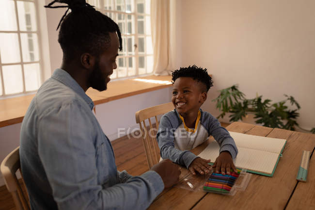 Seitenansicht eines glücklichen afrikanisch-amerikanischen Vaters, der seinem Sohn beim Zeichnen hilft, während er im Esszimmer auf einem Stuhl sitzt — Stockfoto
