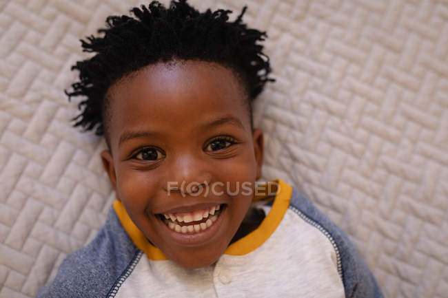 Retrato de um menino afro-americano feliz deitado na cama em casa. Ele está sorrindo e olhando para a câmera — Fotografia de Stock