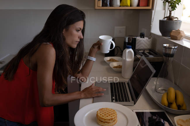 Vista laterale di bella donna mista che lavora sul computer portatile mentre prende il caffè in cucina — Foto stock