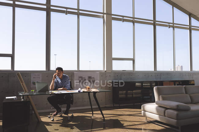 Front view of hardworking Asian male architect sitting at desk and talking on mobile phone in a modern office — Stock Photo