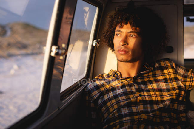 Vista frontal do pensativo homem de corrida mista sentado em uma van campista e olhando para a praia no fundo em um dia ensolarado — Fotografia de Stock