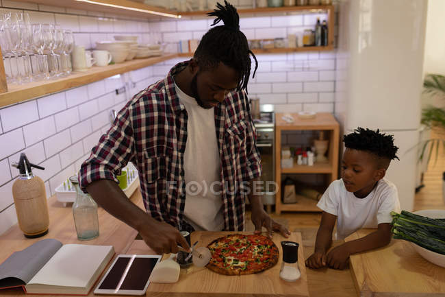 Vista lateral del hijo afroamericano mirando a su padre mientras corta pizza con cortador de pizza en la cocina en casa - foto de stock