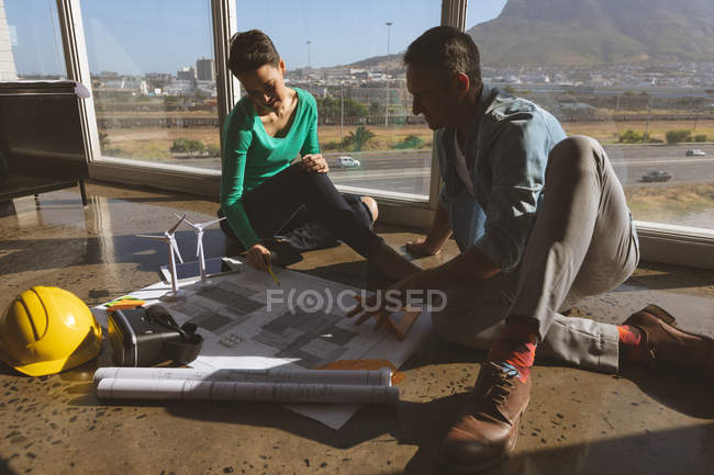 Front view of Caucasian architects sitting on the office ground while they are examining blueprints against window in modern office — Stock Photo