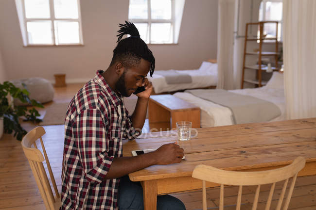 Side view of African-American man talking on mobile phone while using digital tablet at home — Stock Photo
