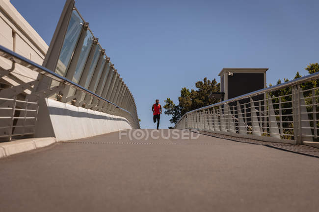 Front view of young African-American fit man with headset running against railing on a sunny day — Stock Photo