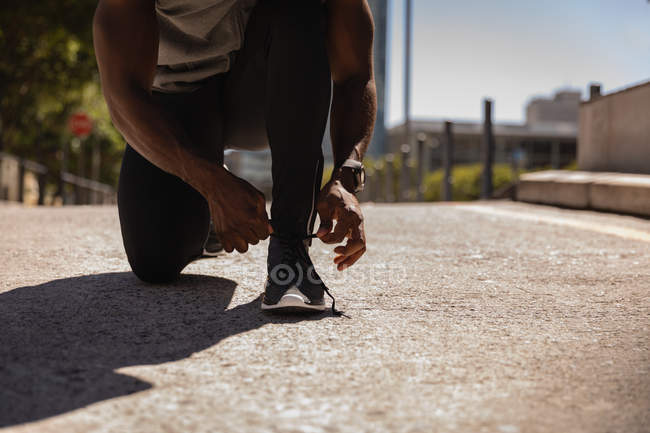 Середина человека, завязывающего шнурки на ботинках, приседая на тротуаре в солнечный день — стоковое фото