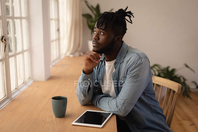 Vista laterale di un uomo afro-americano premuroso che prende un caffè seduto sulla sedia a casa. Sta guardando altrove. — Foto stock