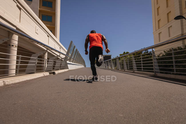 Вид сзади на молодого афроамериканца с наушниками, бегущего против перил в солнечный день — стоковое фото
