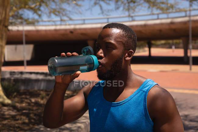 Vista frontal de un joven afroamericano en forma de hombre bebiendo agua mientras está parado en la calle en un día soleado - foto de stock