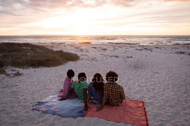 Vista trasera del grupo de amigos multiétnicos sentados en la playa y mirando hacia el océano mientras se pone el sol - foto de stock