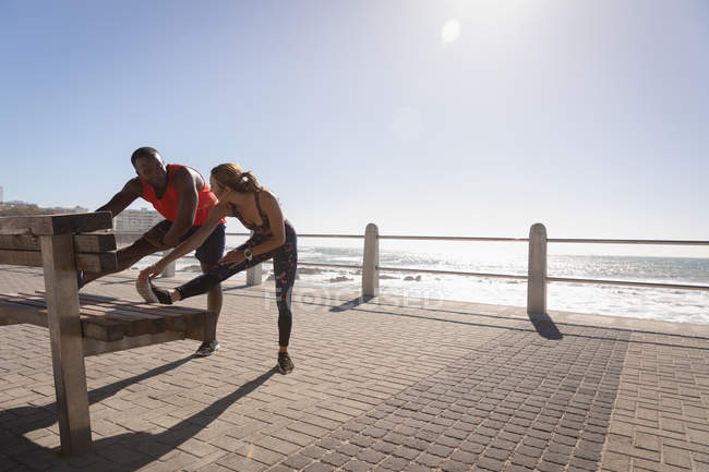 Front view of young multi-ethnic couple doing stretching exercise on bench near beach on a sunny day — Stock Photo