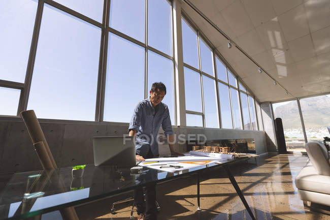 Вид спереди мужчины-архитектора азиатского происхождения, стоящего за столом и смотрящего на синий принт в современном офисе — стоковое фото