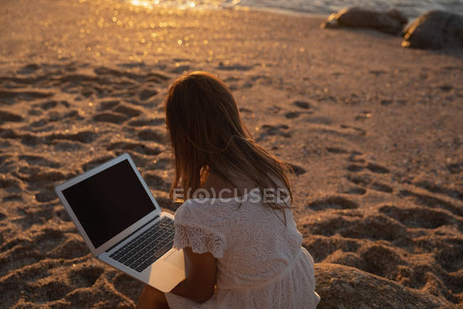 Вид ззаду жінки, використовуючи ноутбук на пляжі під час заходу сонця — стокове фото