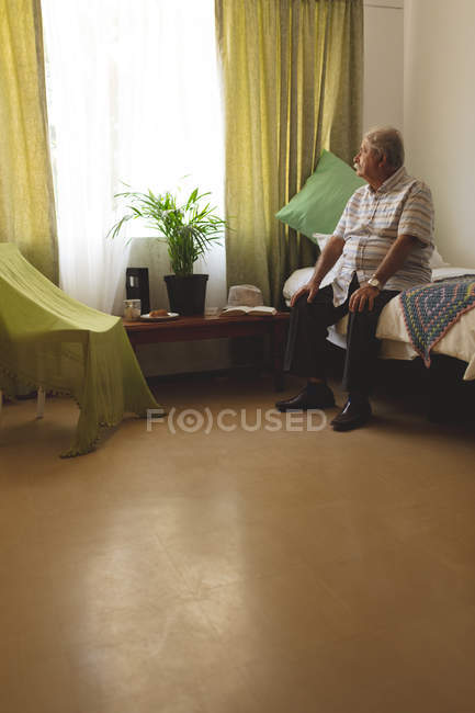 Vista laterale dell'anziano caucasico che guarda fuori dalla finestra mentre è seduto da solo sul letto a casa di cura — Foto stock